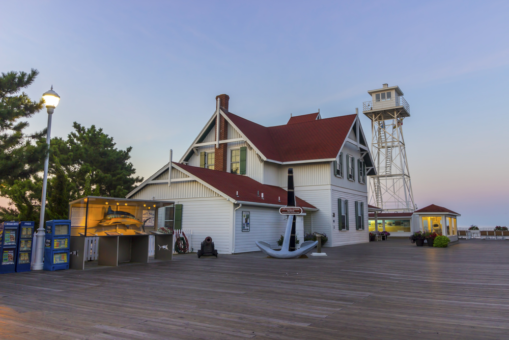 Exterior shot of the Ocean City Life-Saving Museum, a white building with a red roof by the water on the boardwalk.