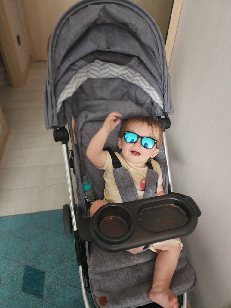 a toddler sits in a stroller smiling and wearing blue reflective sunglasses