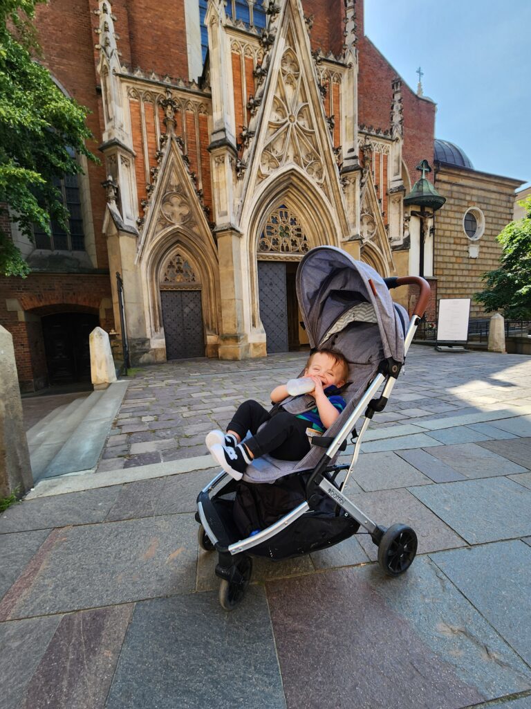 baby sitting in a grey stroller drinking a bottle and smiling with a european church door behind him